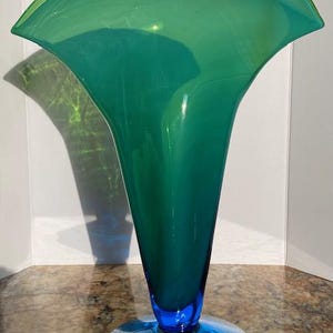 Può includere: Un elegante vaso di vetro con la parte superiore svasata e un corpo conico e sottile. Il vaso passa da un verde-blu intenso a una tonalità verde vibrante. La base è di una tonalità blu a contrasto. Il vaso è alto circa 30 cm.