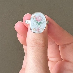 May include: Silver-toned ring with an oval, light blue face featuring a pink rose and floral design. The ring's band has a decorative pattern. The ring is held up by a hand.