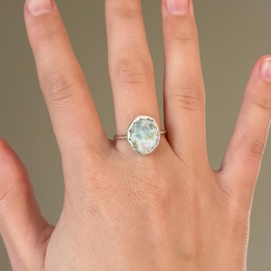 May include: A silver ring with a large, light blue, oval-shaped stone. The stone has a unique, textured appearance with hints of pink and green. The ring is set on a thin silver band, showcasing a delicate and elegant design.