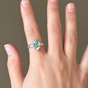 May include: Silver ring featuring an oval-shaped, white enamel centerpiece with a blue floral design and green leaves. The ring's band has a textured pattern. The ring is worn on a finger.