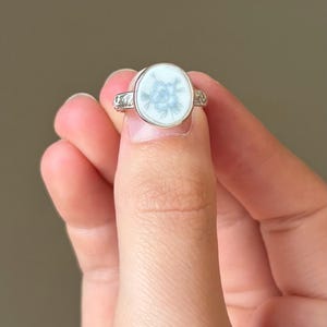 May include: A silver ring with a round, white face featuring a light blue floral design. The ring is held between fingers, showcasing the intricate details of the flower and the ring's band.