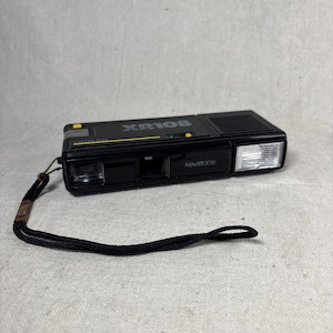 May include: A black Keystone XR108 film camera featuring a rectangular body, a built-in flash unit on the right side, a lens on the left, and a black wrist strap attached to the side. The top of the camera displays the model name XR108.