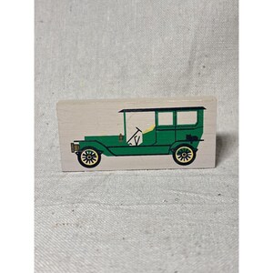 May include: A painted wooden block featuring a green vintage car with black accents. The car has yellow wheels and a black roof, set against a light beige background. The car's design is simple and illustrative.