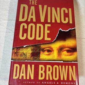 May include: A hardcover book titled The Da Vinci Code by Dan Brown. The red cover features large yellow text and a torn section revealing a close-up of the Mona Lisa's eyes over handwritten script. The text Author of Angels & Demons is at the bottom.
