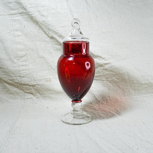 May include: A ruby red glass apothecary jar with a clear glass lid and base. The jar has a bulbous body and a clear, rounded handle on the lid. The base is a clear, circular footed design. The jar is approximately 10 inches tall.