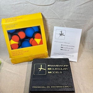 May include: A yellow box of Framework Molecular Model Student Sets, with blue, red, and yellow spheres. A black box with gold lettering and a white instruction manual are also present. A ruler is at the bottom.