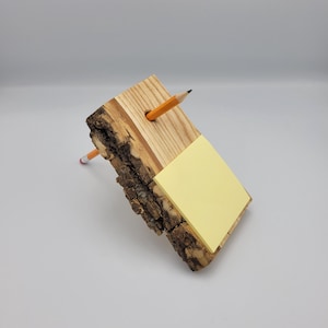 Puede incluir: Un organizador de escritorio de madera con borde de corteza natural, que contiene una pila de notas adhesivas amarillas y dos lápices. El organizador está hecho de madera clara con un agujero del tamaño de un lápiz.