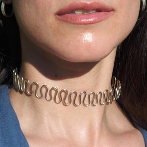 Puede incluir: Un collar tipo gargantilla de metal plateado que presenta un diseño de alambre ondulado o serpentino continuo y repetitivo que rodea el cuello. La pieza tiene un acabado metálico liso y un patrón rítmico constante en toda su longitud.