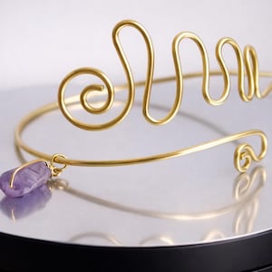 Puede incluir: Brazalete dorado con un diseño de alambre en espiral. Una pequeña gema de amatista púrpura cuelga del brazalete. El brazalete se muestra sobre una superficie reflectante, resaltando sus intrincados detalles y su elegante forma.