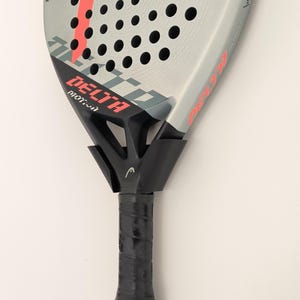 May include: A grey and black padel racket featuring a prominent coral-colored Head logo and the text Delta Motion printed on the face. The racket has a perforated surface, a black handle with a grip, and a black wrist strap attached at the base.