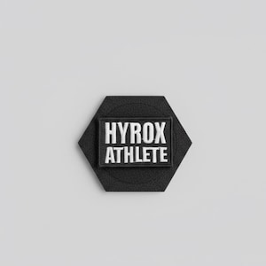 Pu&ograve; includere: Una toppa esagonale nera con le parole "HYROX ATHLETE" ricamate in bianco. La toppa ha una sezione rettangolare al centro con il testo. Lo sfondo &egrave; grigio chiaro.