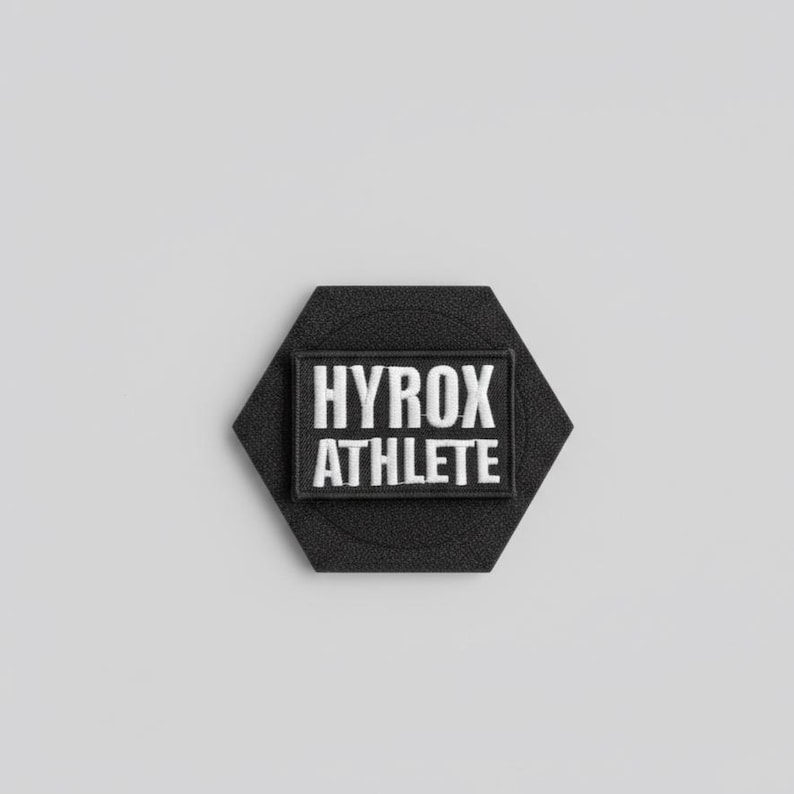 Pu&ograve; includere: Una toppa esagonale nera con le parole "HYROX ATHLETE" ricamate in bianco. La toppa ha una sezione rettangolare al centro con il testo. Lo sfondo &egrave; grigio chiaro.