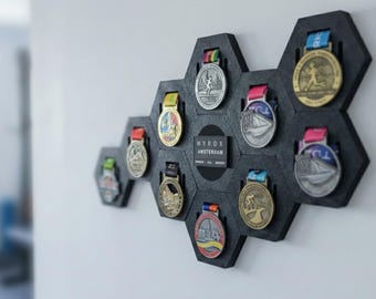 May include: A dark gray, honeycomb-shaped medal display mounted on a white wall. The display holds several medals with colorful ribbons. The medals are of various designs, some with text and images. The display also features a small black plaque with white text that reads "HYROX AMSTERDAM."