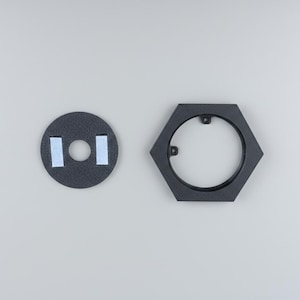 Pu&ograve; includere: Due forme geometriche nere e strutturate su sfondo grigio. Una &egrave; un cerchio con un foro centrale e due strisce adesive. L'altra &egrave; una cornice esagonale nera con un'apertura circolare.