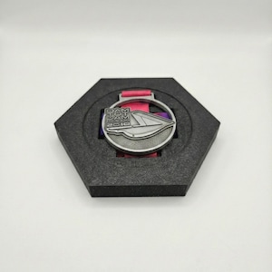 Pu&ograve; includere: Medaglia color argento con la scritta "DAM TOM LOGOS 2016" e un design stilizzato, presentata in un astuccio esagonale nero con un nastro rosa e viola.