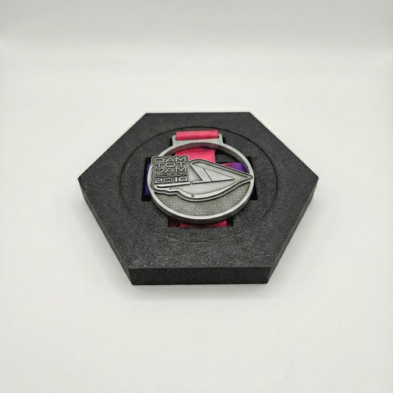 Pu&ograve; includere: Medaglia color argento con la scritta "DAM TOM LOGOS 2016" e un design stilizzato, presentata in un astuccio esagonale nero con un nastro rosa e viola.