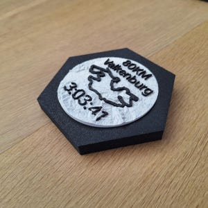 Puede incluir: Un posavasos negro, de forma hexagonal, con un emblema circular blanco. El emblema presenta el texto "80KM Valkenburg" y "3:03:47" en negro. El posavasos probablemente está hecho de un material texturizado.