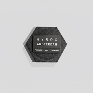 Pu&ograve; includere: Una medaglia da finisher esagonale nera con la scritta "HYROX AMSTERDAM" e "FINISHER NLD 2024/2025" in bianco. La medaglia ha una superficie strutturata ed &egrave; posta su uno sfondo bianco.