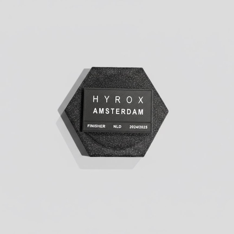 Pu&ograve; includere: Una medaglia da finisher esagonale nera con la scritta "HYROX AMSTERDAM" e "FINISHER NLD 2024/2025" in bianco. La medaglia ha una superficie strutturata ed &egrave; posta su uno sfondo bianco.