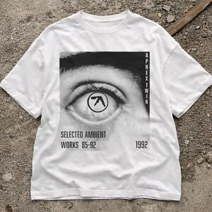 May include: White t-shirt featuring a black and white graphic of an eye with a logo in the iris. The text "Aphex Twin" is printed vertically on the right side, with "SELECTED AMBIENT WORKS 85-92" and "1992" below the eye graphic.