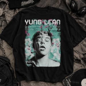 Op de afbeelding: Een zwart t-shirt met korte mouwen en een grafische print van Yung Lean. Het ontwerp bevat de tekst "YUNG LEAN" bovenaan, songtitels zoals "Ginseng Strip 2002," "Kyoto," "Yoshi City," "Ghosttown," "Blinded," en "Creep Creeps" met bloemige accenten.