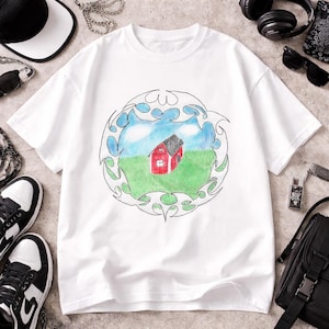 May include: A white short-sleeve t-shirt featuring a central graphic of a red barn in a green field under a blue sky, enclosed within a black-outlined tribal-style circular frame. The design appears to be a hand-drawn illustration.