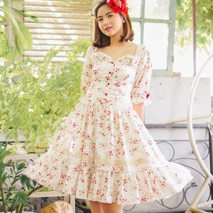 May include: A woman wearing a white dress with a floral pattern and red flowers in her hair. The dress has a sweetheart neckline and a ruffled skirt. She is standing in a garden setting.
