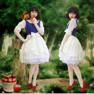May include: Three women dressed as Snow White in a white dress with a blue bodice and white lace trim. They are wearing white gloves and red shoes. The women are standing in a forest setting.