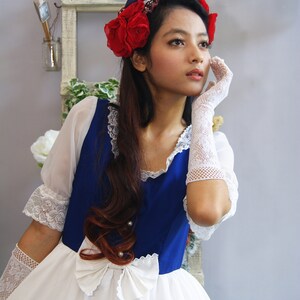 May include: A woman wearing a white dress with a blue bodice and a red flower crown. She is wearing white lace gloves and has her hand on her cheek.