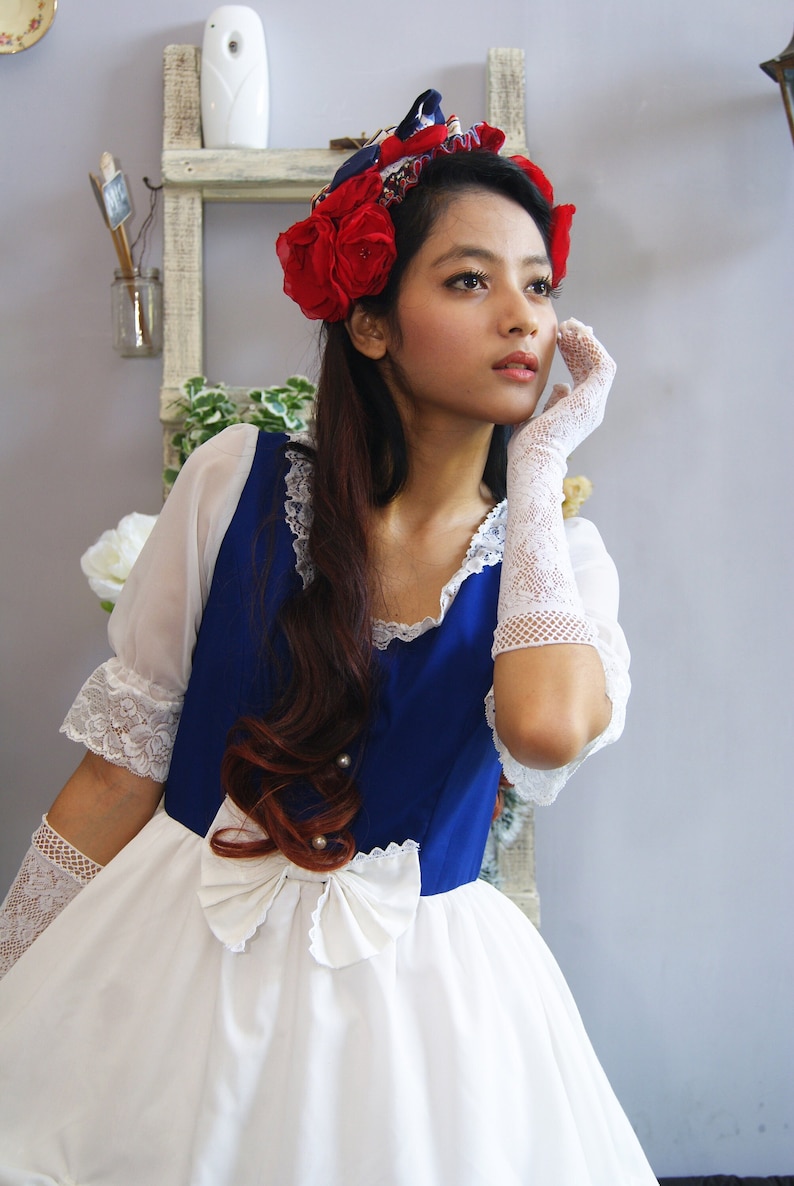 May include: A woman wearing a white dress with a blue bodice and a red flower crown. She is wearing white lace gloves and has her hand on her cheek.