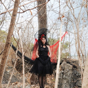 May include: A woman in a black dress with a red flower in her hair stands in a woodland setting. She is wearing a black veil and black gloves. The background is a blurred image of trees and branches.