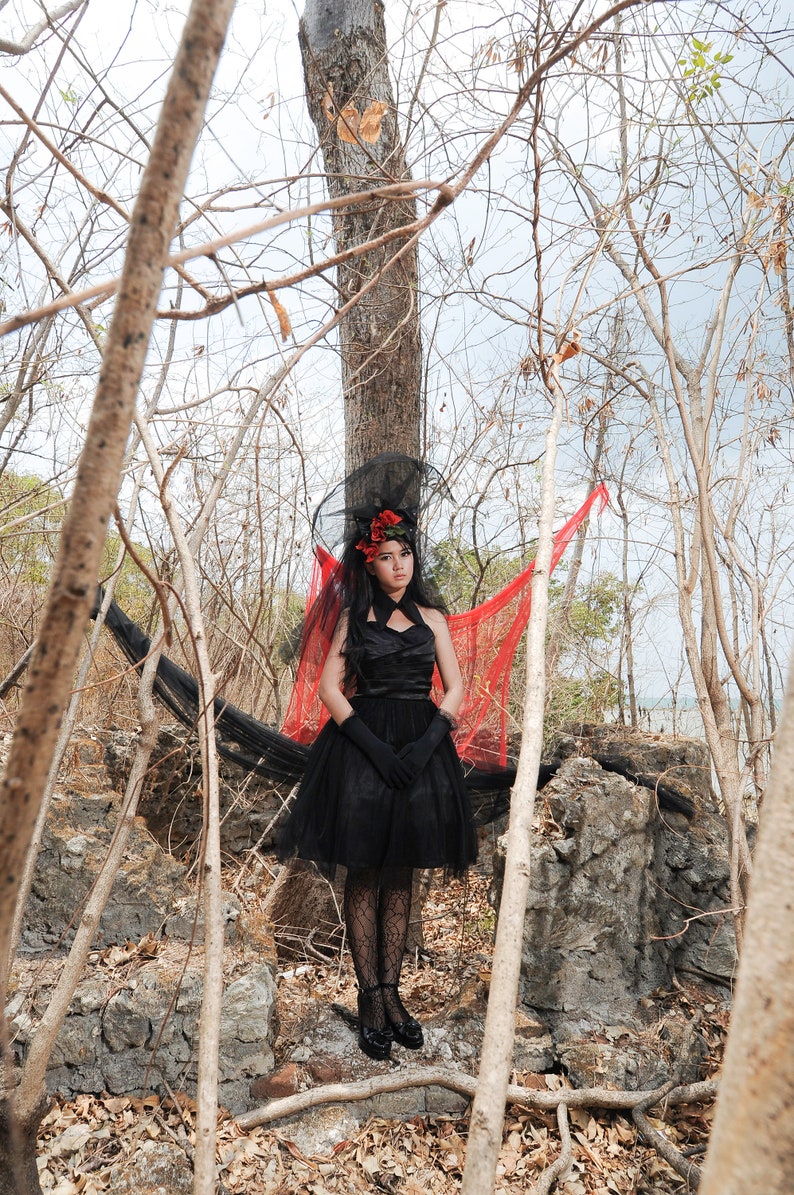 May include: A woman in a black dress with a red flower in her hair stands in a woodland setting. She is wearing a black veil and black gloves. The background is a blurred image of trees and branches.