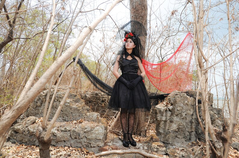 May include: A woman in a black dress with a red and black veil stands in a forest setting. She is wearing black gloves and black shoes. The background is a forest with trees and rocks.