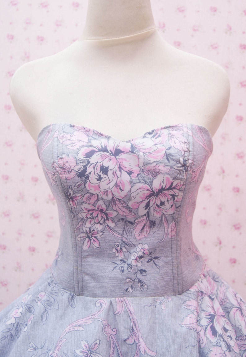 May include: A strapless bodice dress featuring a grey fabric base with a printed pattern of large pink and dark grey floral motifs. The garment has a sweetheart neckline and vertical seam detailing along the front, extending into a full skirt.