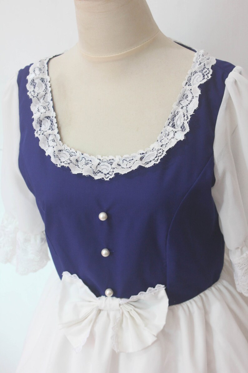 May include: A white dress with a blue bodice and white lace trim. The bodice has pearl buttons and a white bow at the waist.