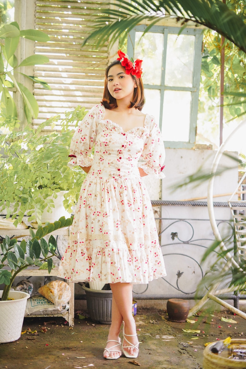 May include: A woman wearing a white dress with a floral pattern and red flowers in her hair. She is standing in front of a white wall with a window and plants.