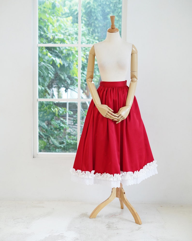 May include: A red skirt with a white lace trim. The skirt is full and has a gathered waist. The skirt is displayed on a mannequin.