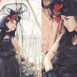 May include: A woman wearing a black dress and veil with red flowers. The veil is made of a sheer fabric and the flowers are arranged in a cluster on the side of her head. The woman is looking down at the camera.