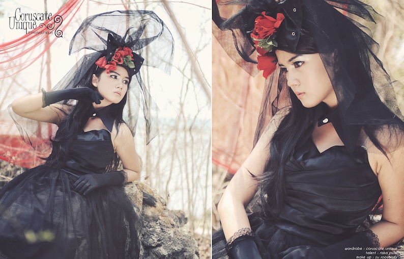 May include: A woman wearing a black dress and veil with red flowers. The veil is made of a sheer fabric and the flowers are arranged in a cluster on the side of her head. The woman is looking down at the camera.