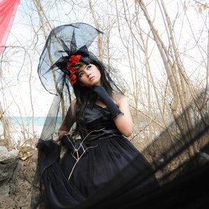 May include: A woman wearing a black dress and a black veil with red flowers. She is standing in a woodland setting with a red fabric in the background.