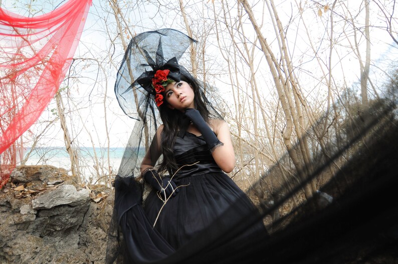 May include: A woman wearing a black dress and a black veil with red flowers. She is standing in a woodland setting with a red fabric in the background.
