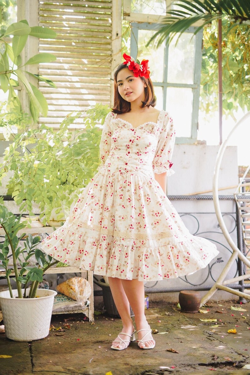 May include: A woman wearing a white dress with a floral pattern and red flowers in her hair. The dress has a sweetheart neckline and a ruffled skirt. She is standing in a garden setting.