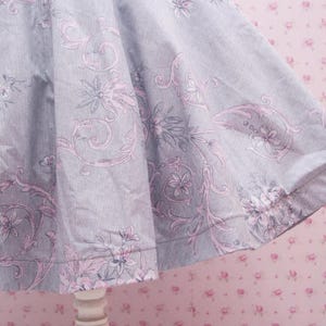 May include: A flared skirt made of light gray fabric featuring a repeating pattern of soft pink floral motifs and intricate swirling vine designs. The garment has a wide, gathered silhouette that creates deep folds and drapes naturally.