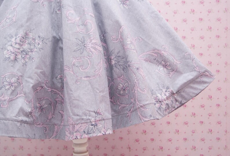 May include: A flared skirt made of light gray fabric featuring a repeating pattern of soft pink floral motifs and intricate swirling vine designs. The garment has a wide, gathered silhouette that creates deep folds and drapes naturally.