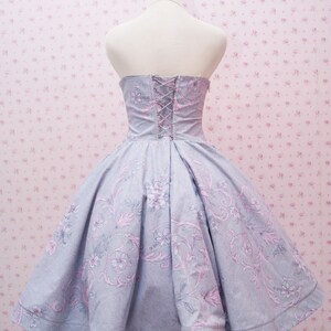 May include: A strapless dress in light blue fabric featuring a pink and grey floral pattern. The bodice includes a lace-up back closure with thin cord lacing, while the skirt has a full, pleated silhouette that flares out from the waistline.