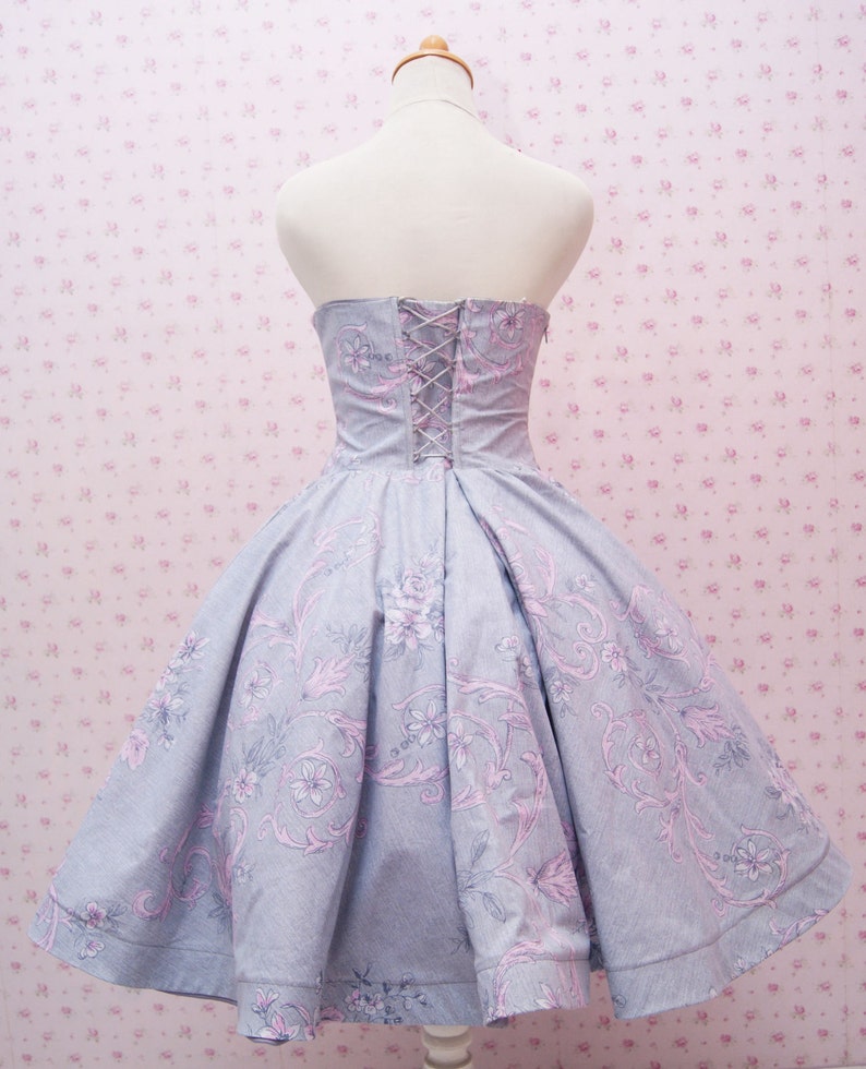 May include: A strapless dress in light blue fabric featuring a pink and grey floral pattern. The bodice includes a lace-up back closure with thin cord lacing, while the skirt has a full, pleated silhouette that flares out from the waistline.