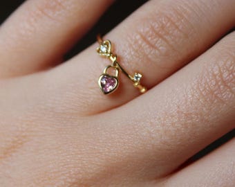 May include: A delicate gold ring featuring a heart-shaped padlock charm with a pink gemstone. The ring also includes a small key and heart-shaped accents, creating a romantic and elegant design. The ring is shown on a finger.
