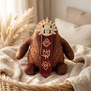 May include: A brown plush toy creature with a sun-shaped headpiece and a maroon scarf. The toy has a patterned design on its body and the scarf features gold symbols. It is sitting in a woven basket, creating a cozy aesthetic.