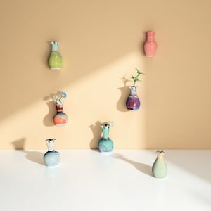Puede incluir: Una colección de pequeños jarrones de cerámica coloridos, algunos con flores y vegetación. Los jarrones son en tonos verdes, azules, rosas y morados, con un acabado texturizado. Están dispuestos contra una pared beige y una superficie blanca.