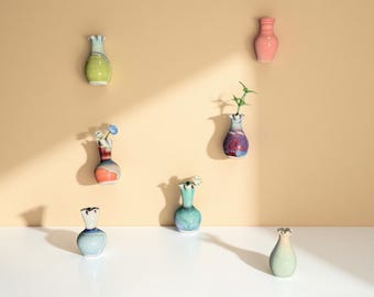 Op de afbeelding: Een verzameling kleine, kleurrijke keramische vazen, sommige met bloemen en groen. De vazen zijn in tinten groen, blauw, roze en paars, met een textuurafwerking. Ze zijn geplaatst tegen een beige muur en een wit oppervlak.